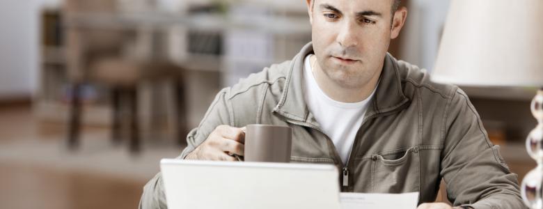 Man sitting in front of a laptop drinking a coffee and holding a piece of paper